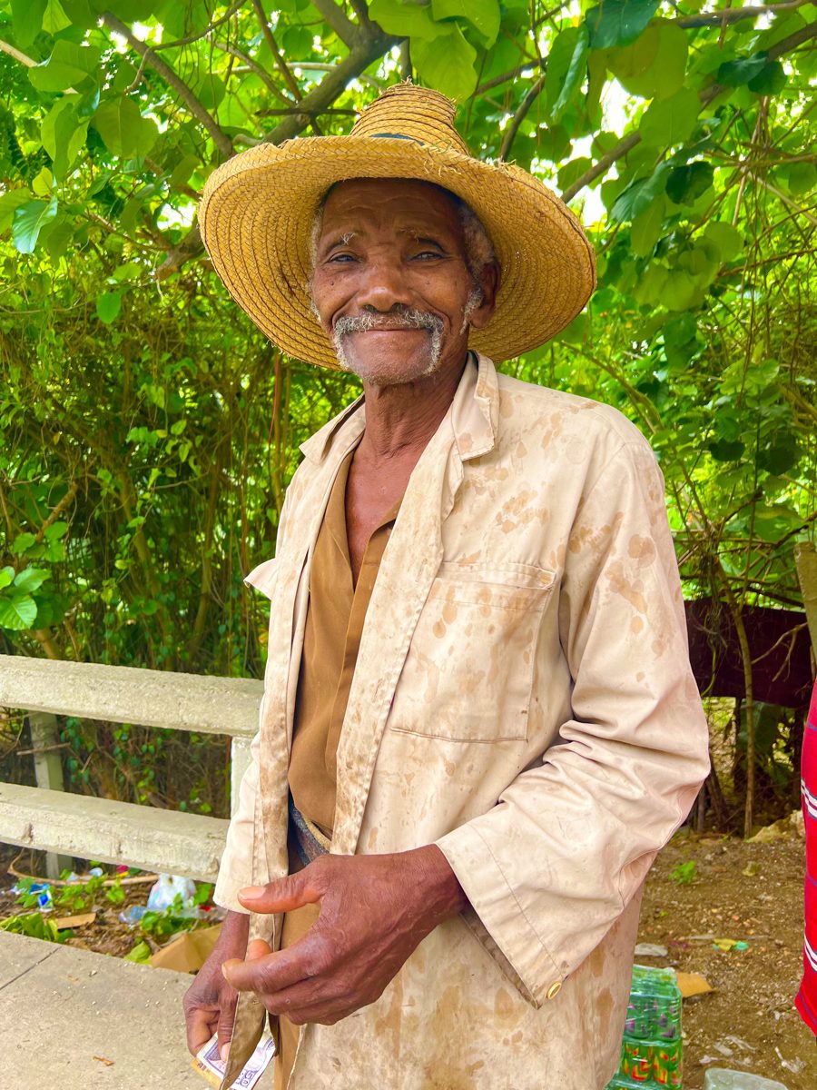 Um agricultor idoso de chapéu de palha sorri em meio à folhagem verde.