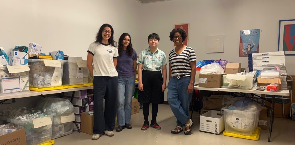 Five volunteers stand beside tables of boxed medical supplies.