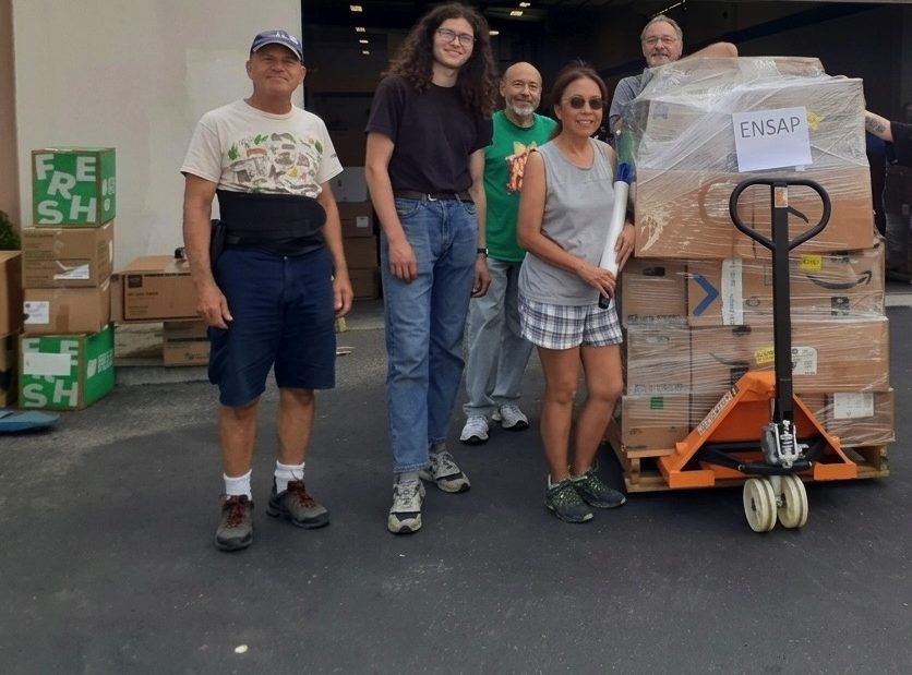 Volunteers stand beside a wrapped pallet of medical supplies outside a warehouse.